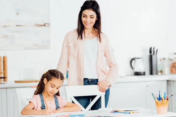 Attraente madre guardando carino figlia disegno a casa — Foto stock