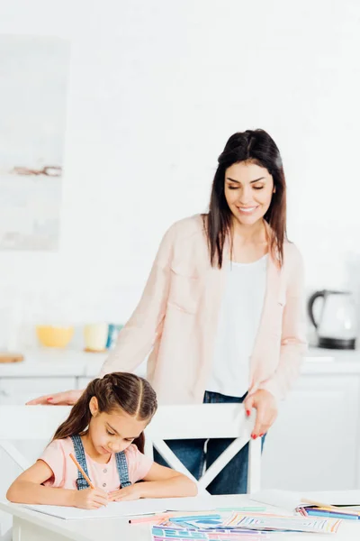 Feliz morena madre mirando lindo hija dibujo en casa - foto de stock