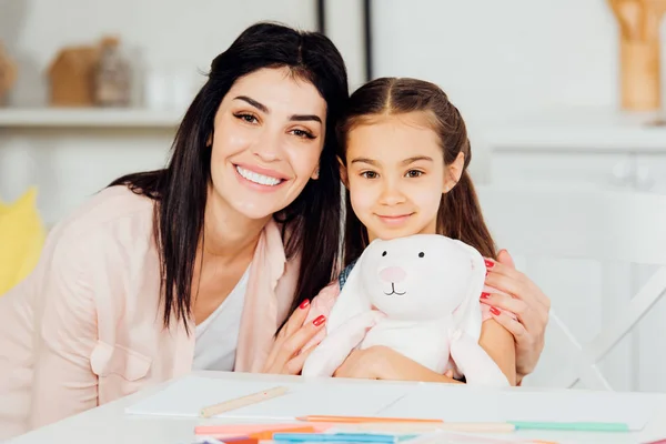 Cheerful daughter holding soft toy near brunette mother and looking at camera — Stock Photo