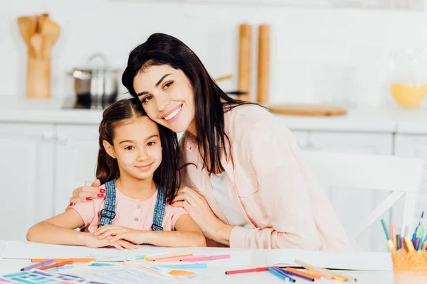 Fröhliche brünette Mutter umarmt glückliche Tochter und schaut in die Kamera — Stockfoto