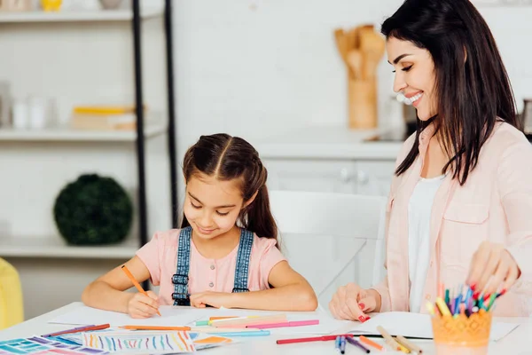 Donna felice guardando allegra figlia disegno e sorridente a casa — Foto stock