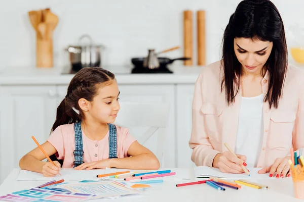 Fille mignonne regardant attrayant brunette mère dessin à la maison — Photo de stock