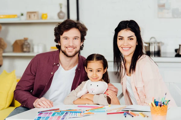 Feliz hombre y mujer sonriendo con linda hija sosteniendo juguete suave y mirando a la cámara - foto de stock