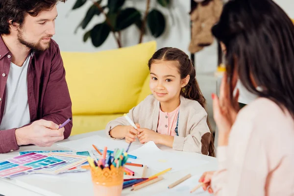 Selektiver Fokus der niedlichen Tochter, die in die Kamera schaut, in der Nähe der brünetten Mutter und des hübschen Vaters — Stockfoto