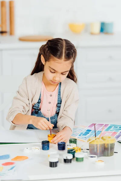 Lindo niño mirando pintura amarilla mientras sostiene el pincel en casa - foto de stock