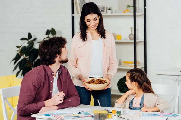 Cheerful mother holding bowl with cookies near husband and daughter — Stock Photo