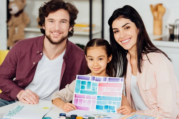 Cheerful kid holding paper with colorful stripes near happy father and mother — Stock Photo