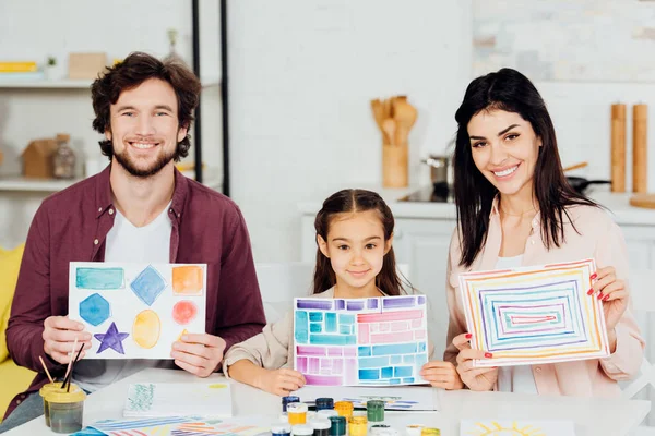 Cheerful kid and happy father and mother holding papers with drawing at home — Stock Photo