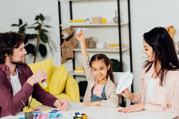 Cheerful kid and happy parents playing with paper planes at home — Stock Photo