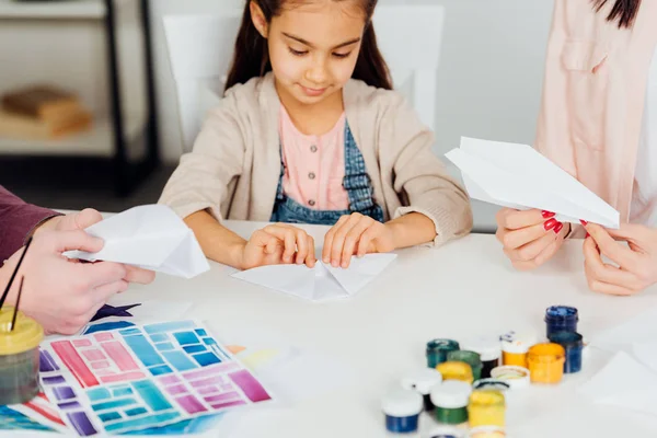 Vista recortada de los padres cerca linda hija con avión de papel en casa - foto de stock