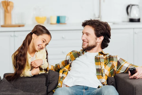 Cheerful father sitting on sofa and holding remote controller near smiling daughter — Stock Photo