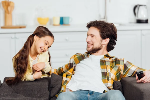 Alegre padre sentado en el sofá y sosteniendo el mando a distancia cerca de sonriente hija - foto de stock