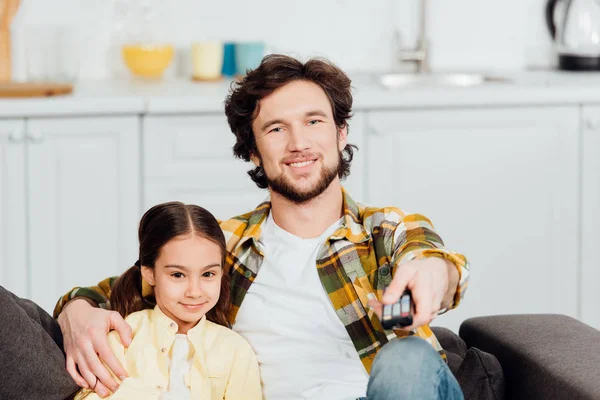 Selective focus of happy father sitting with cute daughter on sofa and holding remote controller — Stock Photo