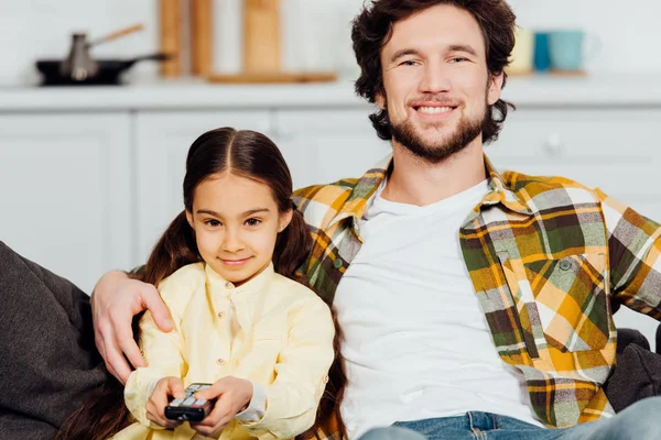 Cute daughter holding remote controller near happy father while sitting on sofa — Stock Photo