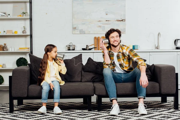 Feliz hija hablando en puede teléfono y alegre padre escuchando mientras está sentado en el sofá - foto de stock