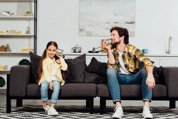 Feliz hija celebración puede teléfono y alegre padre hablando y sentado en el sofá - foto de stock