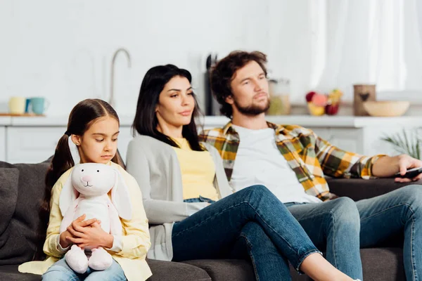 Foco seletivo de criança chateada segurando brinquedo macio perto de pais assistindo TV em casa — Fotografia de Stock