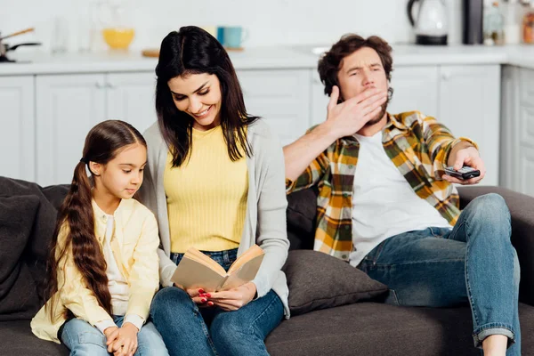 Happy mother reading book to cute daughter near husband yawning on sofa — Stock Photo