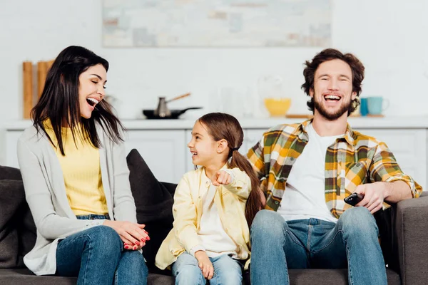 Happy woman looking at daughter pointing with finger near handsome husband with remote controller — Stock Photo