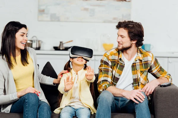 Criança alegre gesto enquanto vestindo fone de ouvido realidade virtual perto de pais felizes em casa — Fotografia de Stock