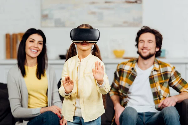 Foco seletivo de criança bonito gesto enquanto vestindo fone de ouvido realidade virtual perto de pais felizes em casa — Fotografia de Stock