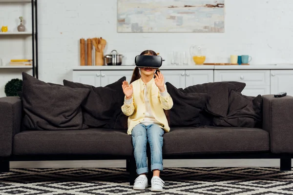 Garoto bonito gesticulando enquanto vestindo fone de ouvido realidade virtual em casa — Fotografia de Stock