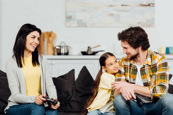 Lindo y feliz niño abrazando padre cerca de madre sosteniendo joystick en casa - foto de stock