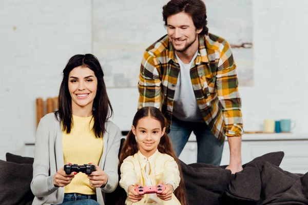 Feliz hombre mirando alegre esposa y niño jugando video juego en casa - foto de stock