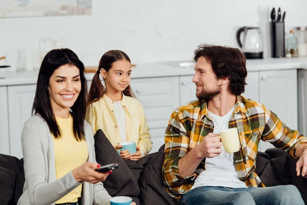 Happy woman holding remote controller near husband and daughter with cups — Stock Photo