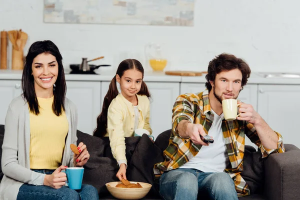 Homme heureux tenant télécommande près de la femme et la fille avec des tasses — Photo de stock