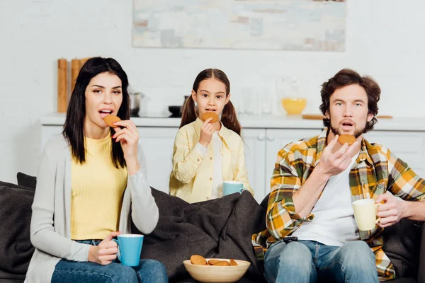 Famille heureuse tenant des tasses avec des boissons tout en mangeant des cookies et en regardant le film — Photo de stock