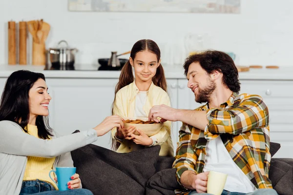 Pais felizes tomando biscoitos perto de filha bonito enquanto segurando copos — Fotografia de Stock