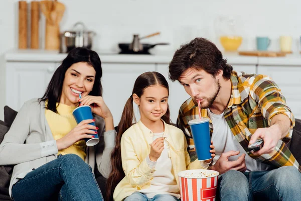 Handsome man drinking soda and holding remote controller near cute daughter and wife — Stock Photo