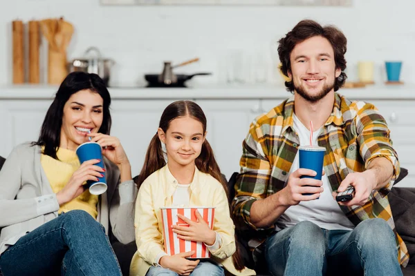 Padres felices sosteniendo vasos desechables cerca de hija alegre con cubo de palomitas de maíz - foto de stock