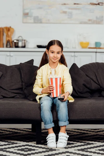 Щаслива дитина тримає пульт дистанційного керування, сидячи на дивані з відром попкорна — стокове фото