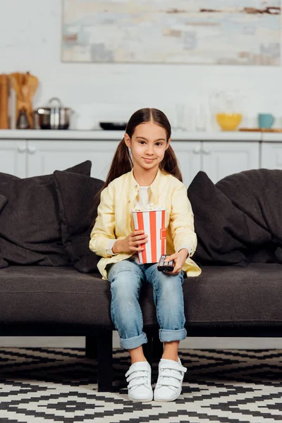 Niño feliz sosteniendo el mando a distancia mientras está sentado en el sofá con cubo de palomitas de maíz - foto de stock