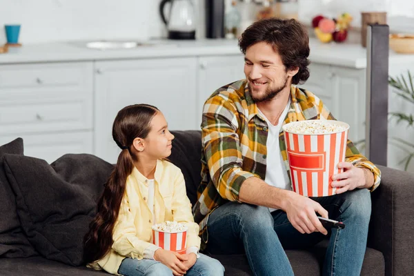 Happy man looking at cute daughter while holding bucket of popcorn and remote controller — Stock Photo