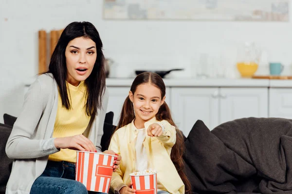 Happy kid pointing with finger while watching movie near surprised mother — Stock Photo