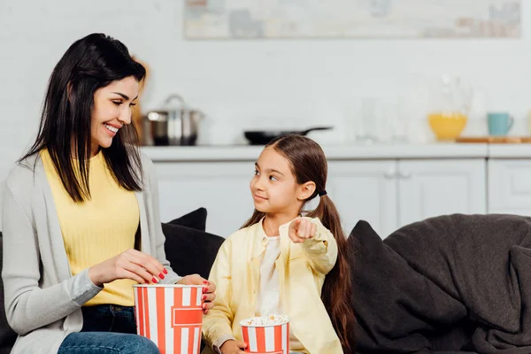 Happy kid pointing with finger and looking at mother holding bucket of popcorn — Stock Photo