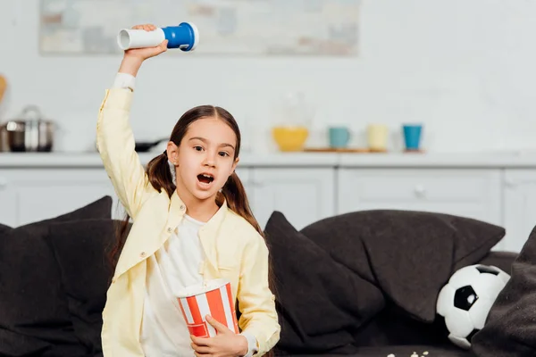 Cute child watching championship and holding bucket of popcorn while screaming and gesturing at home — Stock Photo