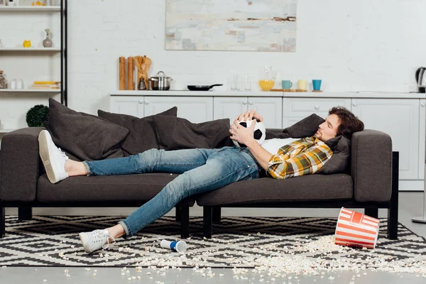Homme fatigué tenant le football et dormant sur le canapé près du seau et du maïs soufflé sur le sol — Photo de stock