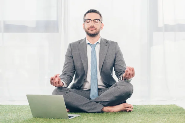 Hombre de negocios sentado en la alfombra de hierba cerca de la computadora portátil y meditando en Lotus Pose - foto de stock