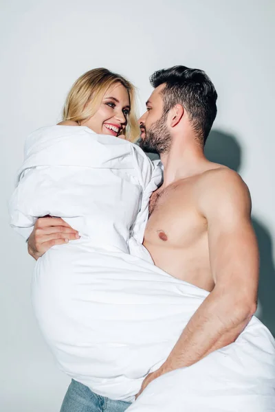 Bonito sem camisa homem segurando no braços feliz loira mulher no cobertor no branco — Fotografia de Stock