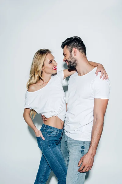 Heureuse femme blonde debout avec la main dans la poche et étreignant homme barbu joyeux sur blanc — Photo de stock