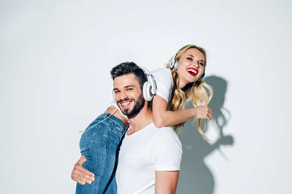 Hombre feliz en auriculares sosteniendo en brazos alegre chica rubia en blanco - foto de stock