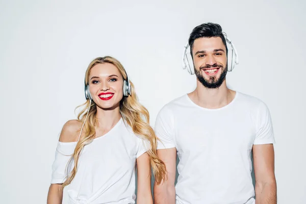 Cheerful bearded man and happy blonde girl looking at camera while listening music in headphones on white — Stock Photo