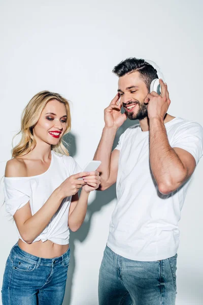 Весела дівчина тримає смартфон біля бородатого чоловіка в навушниках на білому — стокове фото