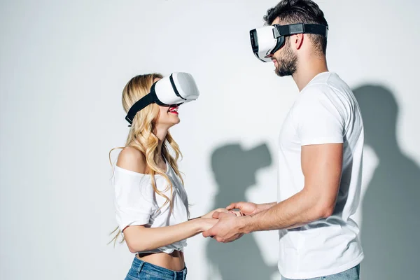 Hombre feliz cogido de la mano con la mujer rubia mientras usa auriculares de realidad virtual en blanco - foto de stock