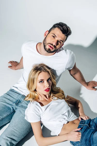 Vue aérienne du bel homme barbu assis et regardant la caméra près de jolie fille blonde avec les mains dans les poches sur blanc — Photo de stock