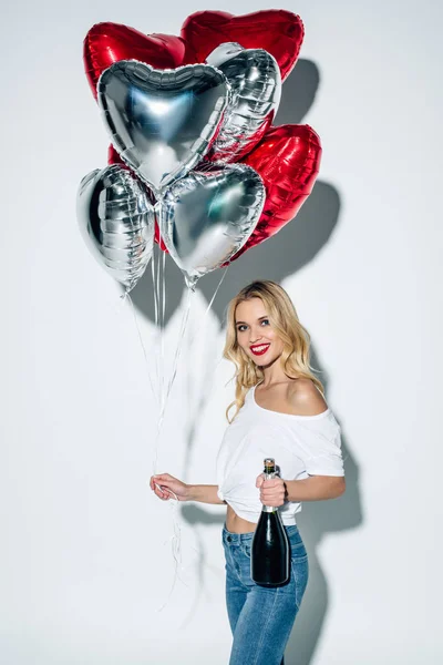 Chica rubia feliz sosteniendo globos y botella de champán mientras está de pie en blanco - foto de stock